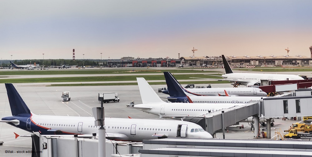 The image shows a busy airport tarmac with multiple airplanes parked at gates. Various aircraft of different sizes and colors are visible, along with ground support vehicles near the planes. The background features a terminal building and additional planes in the distance.
