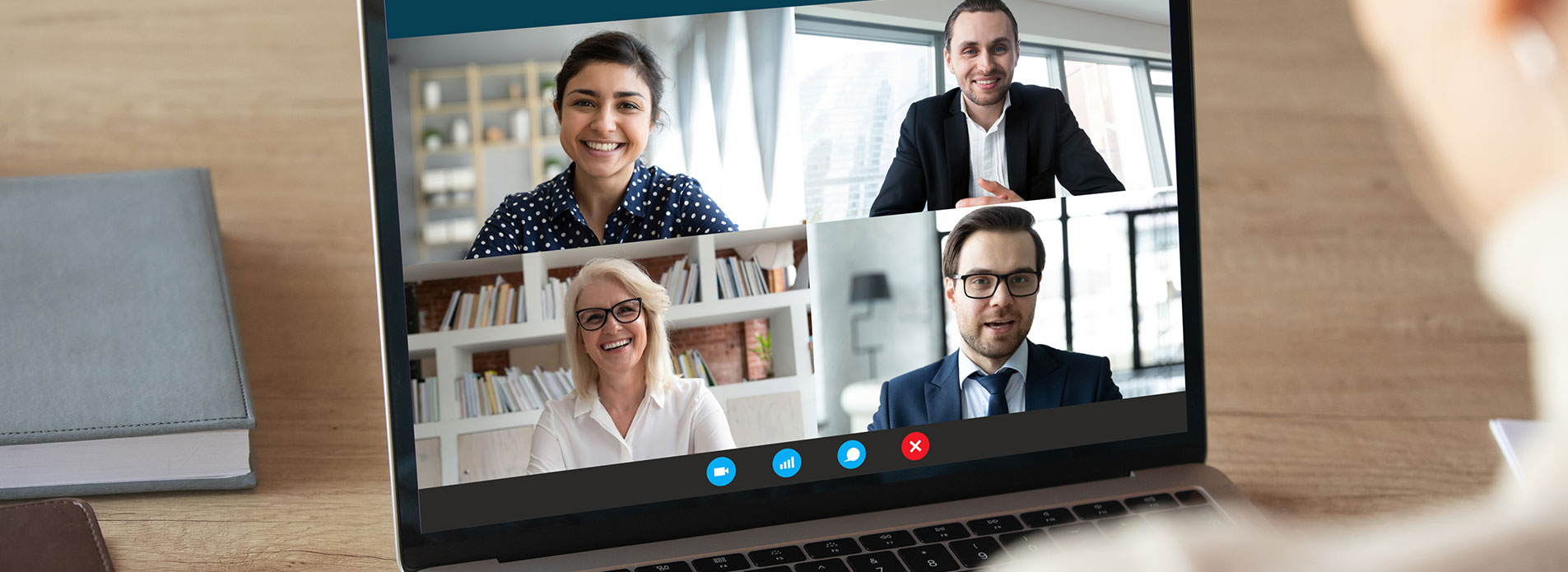 Eine Videokonferenz mit vier Personen auf einem Laptop-Bildschirm. Die Teilnehmer lächeln und scheinen miteinander zu sprechen. Im Hintergrund sind Bücherregale und Fenster zu sehen. Der Tisch ist aus Holz und es liegt ein Buch darauf.