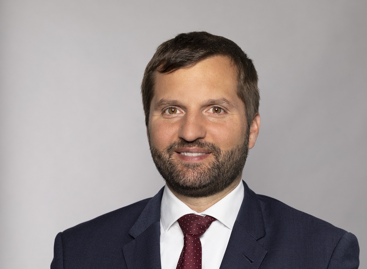 A man with a short beard and brown hair smiles at the camera. He is wearing a dark suit with a white shirt and a maroon tie. The background is a light gray, emphasizing his professional appearance.