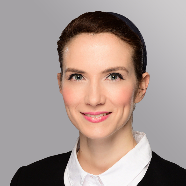 Ein Porträt einer Frau mit schmalem Gesicht, strahlenden grünen Augen und einem freundlichen Lächeln. Sie trägt ein schwarzes Oberteil und eine weiße Bluse. Ihr Haar ist zurückgebunden und sie hat einen schlichten Haargummi im Haar. Der Hintergrund ist graublau.