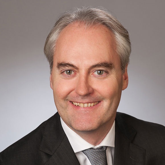 A smiling man with short, light gray hair and light eyes is wearing a dark suit and white shirt with a tie. He appears professional and approachable, set against a simple gray background.