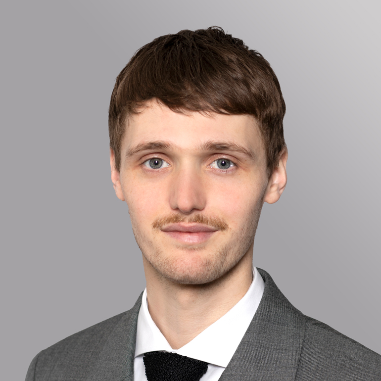 Porträt eines jungen Mannes mit kurzen, braunen Haaren und einem schmalen Schnurrbart. Er trägt einen grauen Anzug mit einer weißen Hemd und einer schwarzen Krawatte. Im Hintergrund ist eine graue, einfarbige Fläche. Der Mann lächelt sanft und schaut direkt in die Kamera.