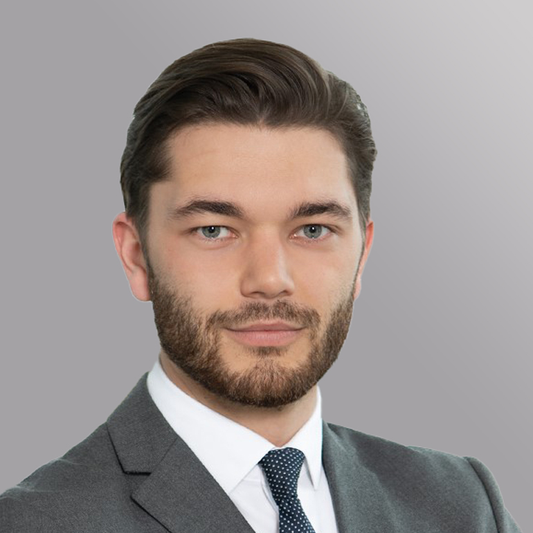 Ein junger Mann mit einem kurzen, gepflegten Bart und braunem Haar trägt einen grauen Anzug und ein weißes Hemd mit einer blauen Krawatte. Er blickt direkt in die Kamera vor einem grauen Hintergrund.