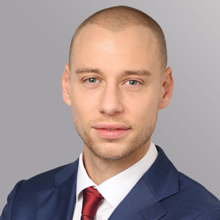 Ein junger Mann mit kurzem, blondem Haar und einem gepflegten Bart. Er trägt einen dunkelblauen Anzug mit einem weißen Hemd und einer roten Krawatte. Der Hintergrund ist grau und unauffällig, um den Fokus auf sein Gesicht zu lenken. Sein Ausdruck ist freundlich und professionell.