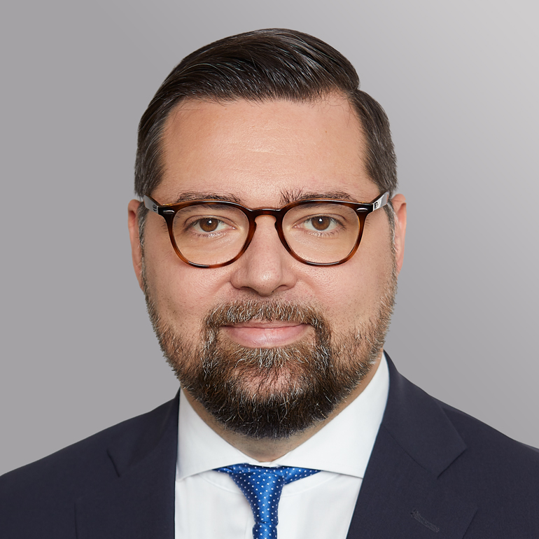Portrait eines Mannes mit Brille, kurzem Haar und Bart. Er trägt einen Anzug mit einem weißen Hemd und einer blauen Krawatte. Der Hintergrund ist einfarbig grau. Der Mann lächelt leicht, was eine freundliche und professionelle Ausstrahlung vermittelt.