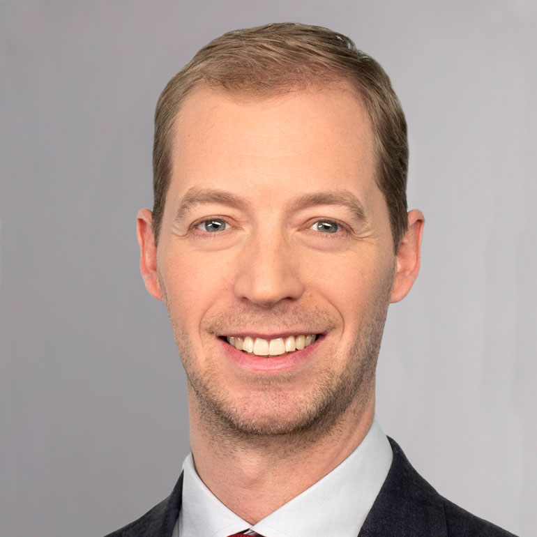 Ein lächelnder Mann mit kurzer, blonder Haaren und einem gepflegten Bart trägt einen dunklen Anzug und eine helle Krawatte. Der Hintergrund ist neutral und einfarbig.