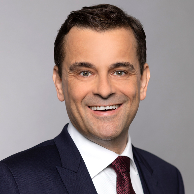 Ein lächelnder Mann mit dunkelbraunem Haar trägt einen dunklen Anzug und eine rote Krawatte. Im Hintergrund ist eine helle, einfarbige Fläche zu sehen. Der Ausdruck des Mannes vermittelt Freundlichkeit und Professionalität.