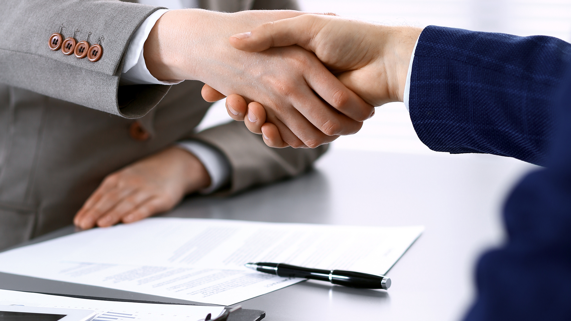 Zwei Personen schütteln sich die Hände über einem Schreibtisch. Auf dem Tisch liegen Dokumente, und ein Kugelschreiber ist sichtbar. Die Szene vermittelt ein Gefühl von Einigung oder Abschluss.