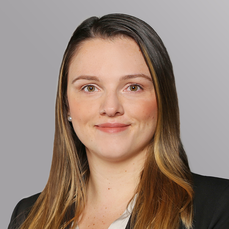 Porträt einer jungen Frau mit langen, glatten, hellen Haaren und einem freundlichen Gesichtsausdruck. Sie trägt eine schwarze Jacke über einem weißen Oberteil. Der Hintergrund ist in einem sanften Grauton gehalten.