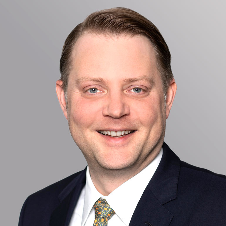 Portrait eines lächelnden Mannes mit kurzen, glatten Haaren. Er trägt einen dunklen Anzug und eine gemusterte Krawatte. Der Hintergrund ist grau und neutral gehalten, wodurch der Fokus auf dem Gesicht des Mannes liegt.