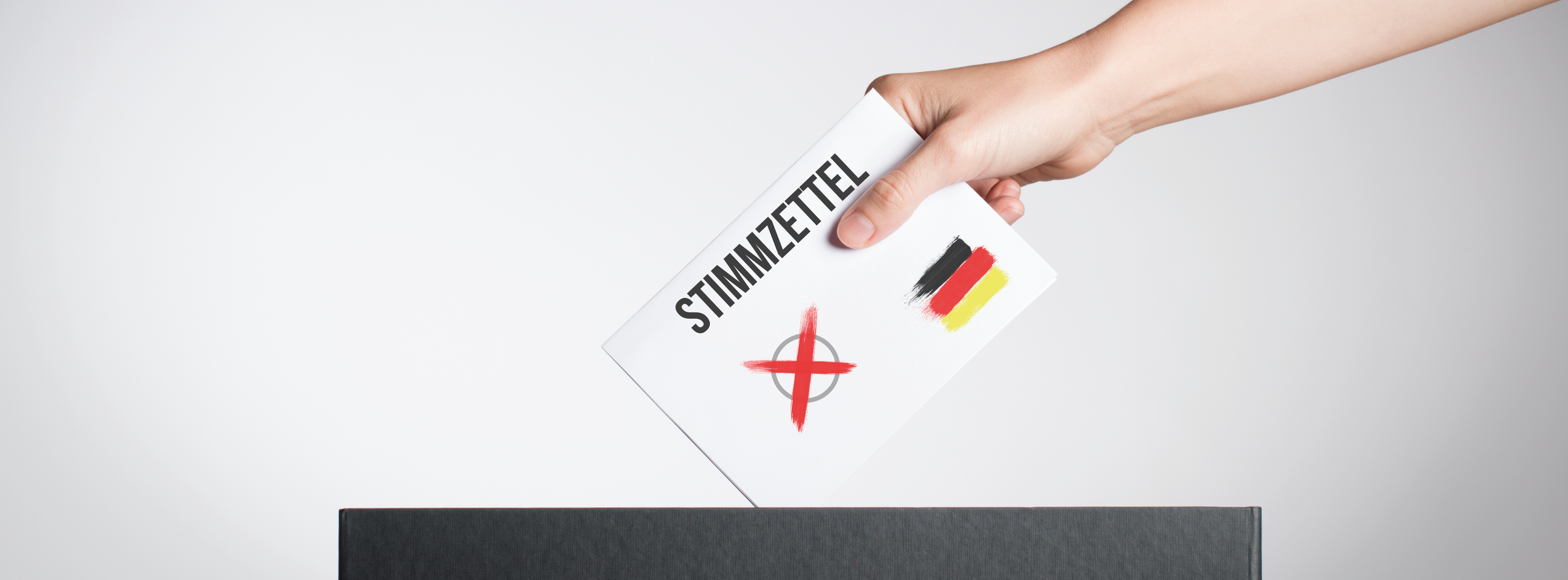 Eine Hand hält einen Stimmzettel mit dem Wort "STIMMZETTEL" und einem roten Kreuz. Der Zettel wird in eine schwarze Wahlurne eingeworfen. Im Hintergrund sind einfarbige Flächen sichtbar. Es symbolisiert die Teilnahme an einer Wahl in Deutschland.
