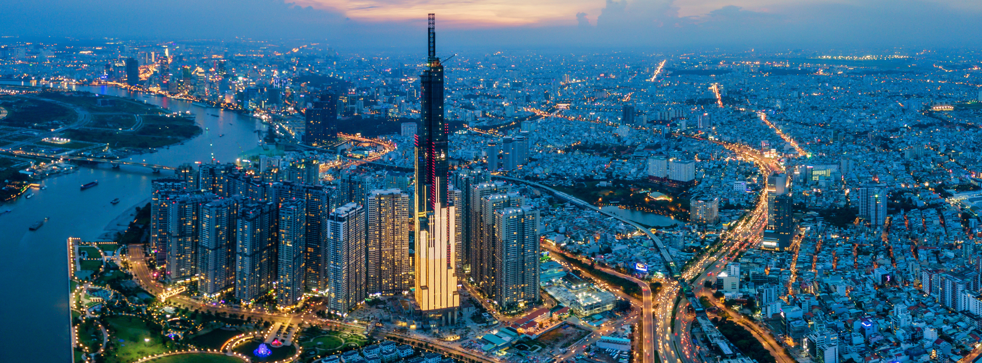 Die Aufnahme zeigt die beeindruckende Skyline von Ho-Chi-Minh-Stadt bei Dämmerung. Im Mittelpunkt steht der Landmark 81, das höchste Gebäude Vietnams, umgeben von weiteren Hochhäusern und einem geschäftigen Stadtbild, das von Lichtern erleuchtet wird. Ein Fluss schlängelt sich durch die Stadt.
