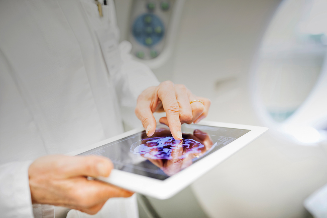 Ein Arzt in einem weißen Kittel hält ein Tablet, auf dem ein Bild eines Gehirns zu sehen ist. Er zeigt mit dem Finger auf das Bild, während er sich in einem klinischen Umfeld befindet.