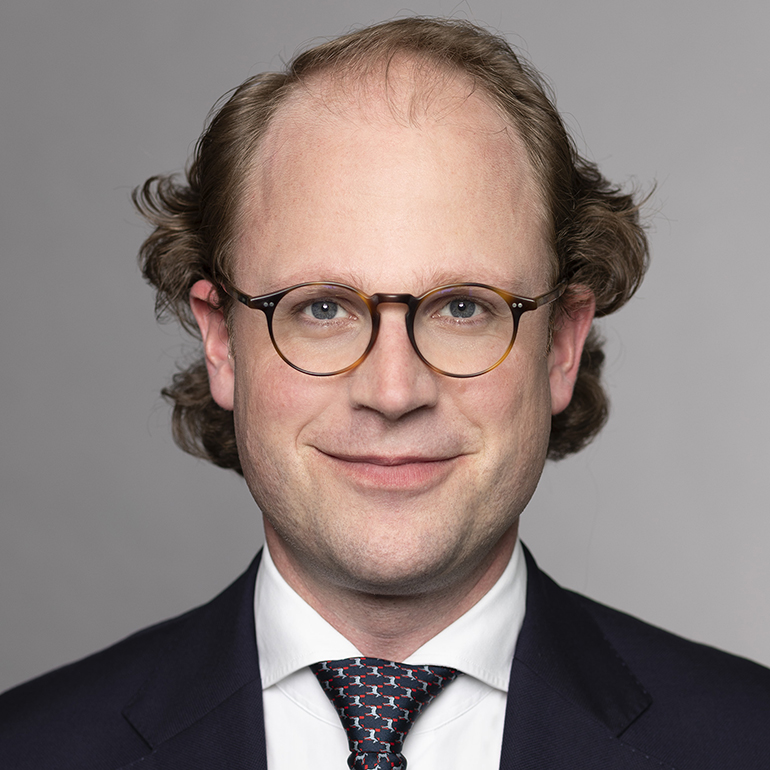 Porträt eines Mannes mit lockigem Haar und einer Brille. Er trägt einen dunklen Anzug und ein weißes Hemd mit einer gemusterten Krawatte. Der Hintergrund ist grau und neutral, um den Fokus auf das Gesicht zu lenken. Der Mann lächelt freundlich.