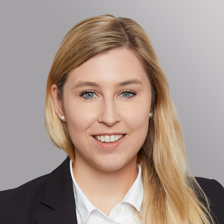 Eine junge Frau mit blonden Haaren und einer leichten Welle lächelt in die Kamera. Sie trägt einen schwarzen Blazer und ein weißes Hemd. Der Hintergrund ist in sanften Grautönen gehalten.