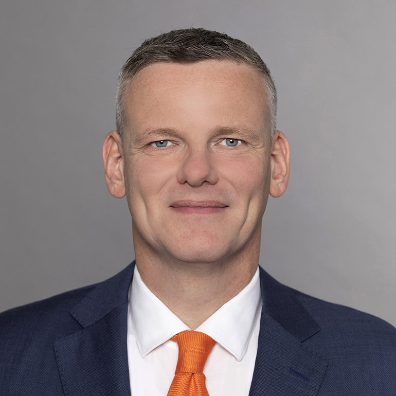 Das Bild zeigt einen Mann mit kurzem, grauem Haar und einem freundlichen Lächeln. Er trägt einen dunkelblauen Anzug und ein weißes Hemd mit einer auffälligen orangefarbenen Krawatte. Der Hintergrund ist grau und schlicht gestaltet.