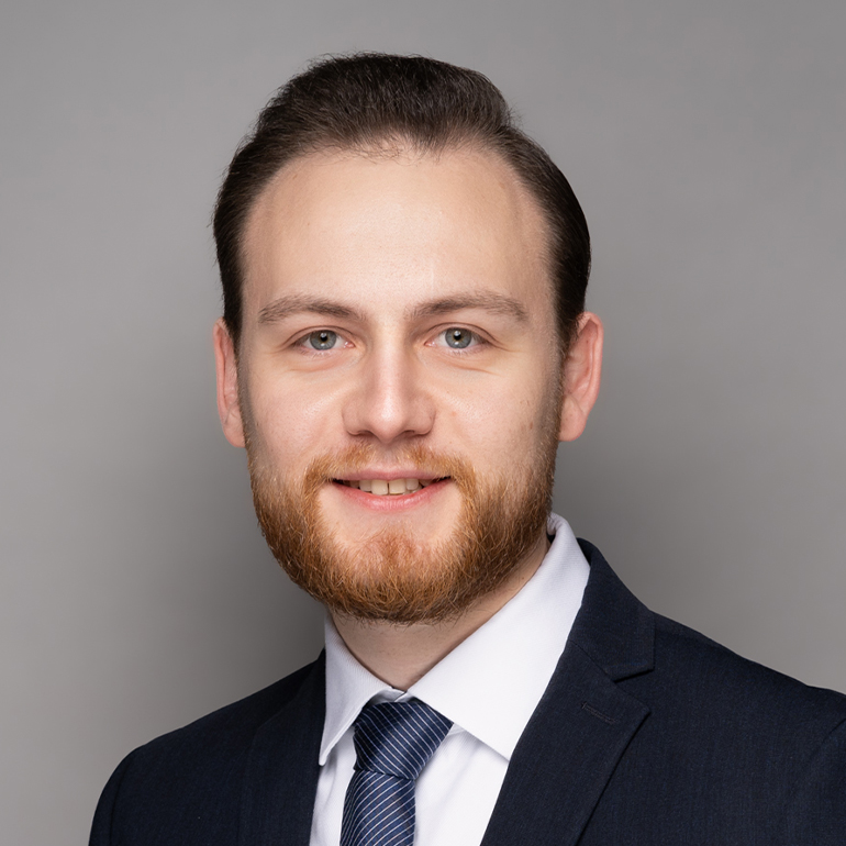 Porträt eines lächelnden Mannes mit Bart, der einen Anzug und eine Krawatte trägt. Der Hintergrund ist grau. Der Mann hat kurzes, dunkles Haar und schaut direkt in die Kamera.