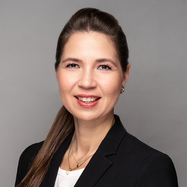 Porträt einer Frau mit langen, braunen Haaren und einem freundlichen Lächeln. Sie trägt einen schwarzen Blazer über einer weißen Bluse. Der Hintergrund ist grau, was einen professionellen Eindruck vermittelt. Die Frau blickt direkt in die Kamera.