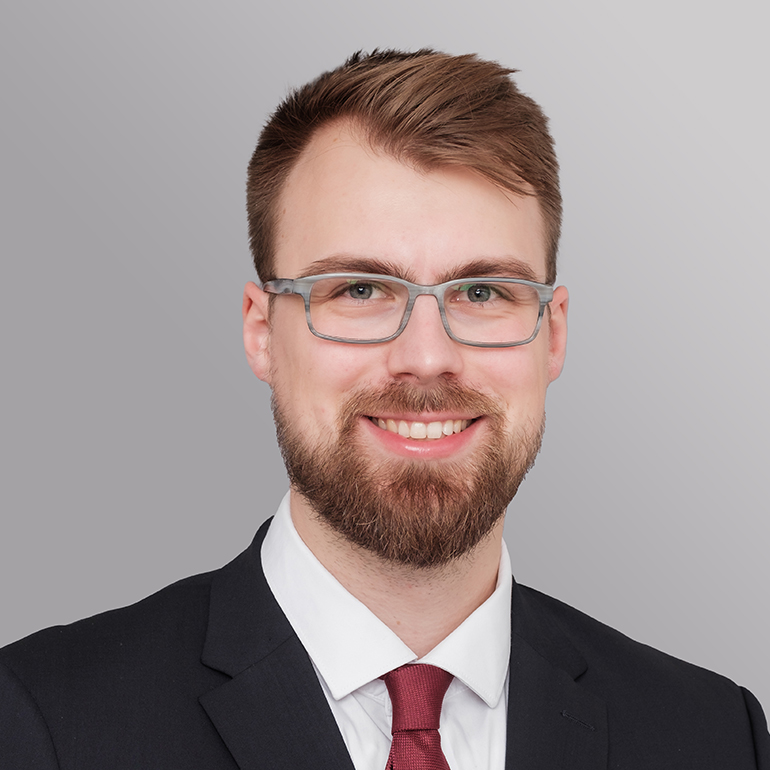 Porträt eines jungen Mannes mit Brille und Bart. Er trägt einen schwarzen Anzug und eine weiße Hemd mit roter Krawatte. Der Hintergrund ist grau. Der Mann lächelt freundlich.