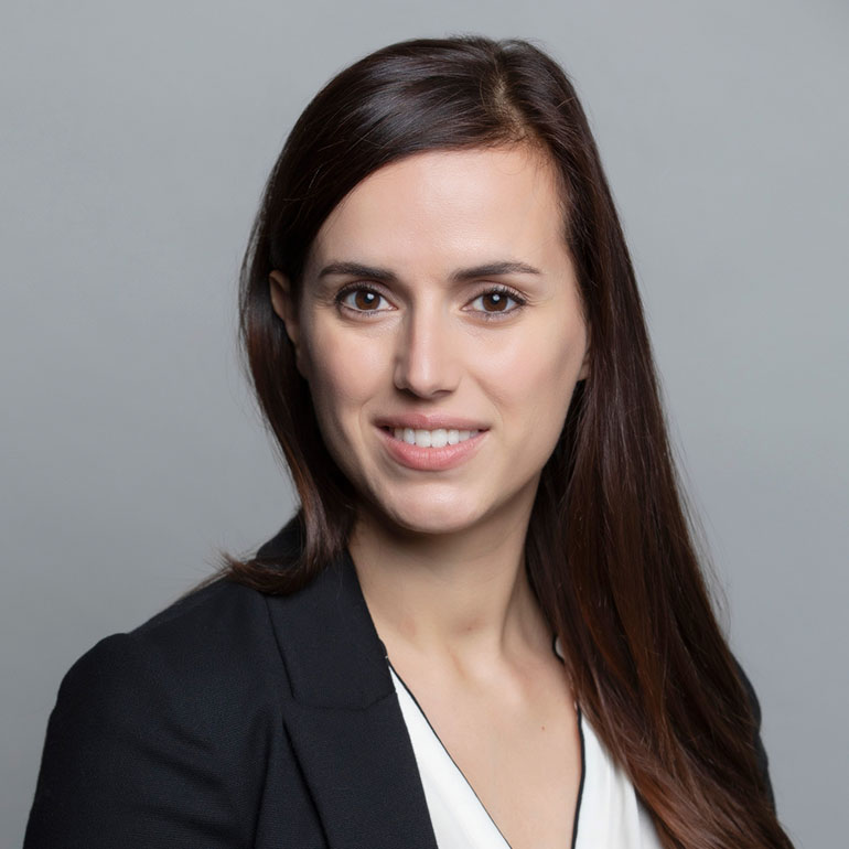 Ein Porträt einer Frau mit langen, braunen Haaren, die in einem schwarzen Blazer und einem weißen Oberteil gekleidet ist. Sie steht vor einem grauen Hintergrund und lächelt freundlich in die Kamera.