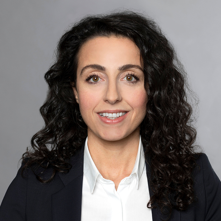 Porträt einer Frau mit langen, lockigen, dunklen Haaren. Sie trägt ein schwarzes Blazerjackett und ein weißes Hemd. Die Frau lächelt freundlich in die Kamera, vor einem neutralen grauen Hintergrund.