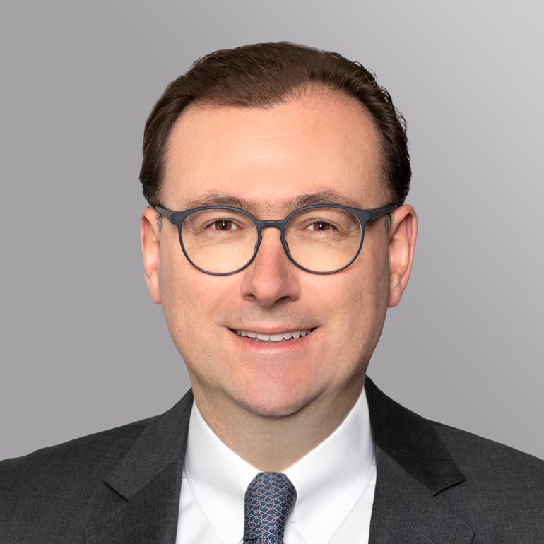 Ein lächelnder Mann mit Brille trägt einen Anzug und ein gemustertes Hemd. Er hat kurze, dunkle Haare und steht vor einem einfarbigen Hintergrund.