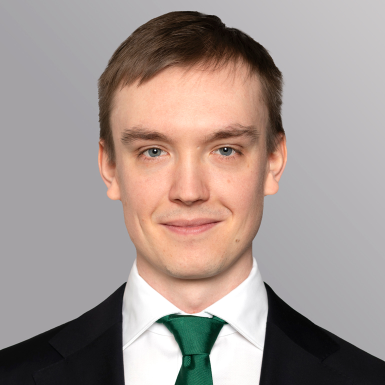 Ein junger Mann mit kurzen, blonden Haaren trägt einen schwarzen Anzug und eine grüne Krawatte. Er schaut direkt in die Kamera und lächelt freundlich. Der Hintergrund ist grau und sorgt für einen neutralen Kontrast zu seinem Outfit.