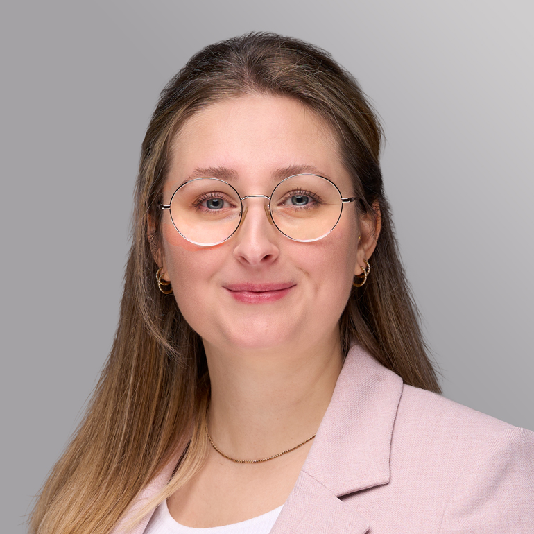 Porträt einer Frau mit langen, blonden Haaren und Brille. Sie trägt einen blauen Oberteil und einen hellen Blazer. Der Hintergrund ist grau. Die Frau lächelt freundlich und schaut direkt in die Kamera.