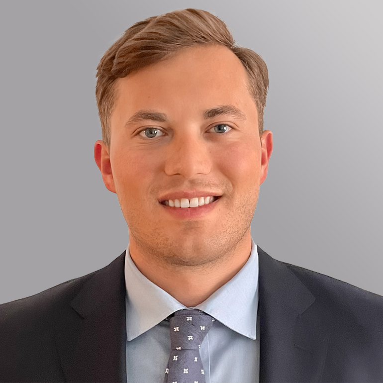 Porträt eines lächelnden Mannes mit kurzen, hellen Haaren, der einen dunklen Anzug und ein hellblaues Hemd trägt. Er hat eine graue Krawatte mit einem Muster und steht vor einem grauen Hintergrund.