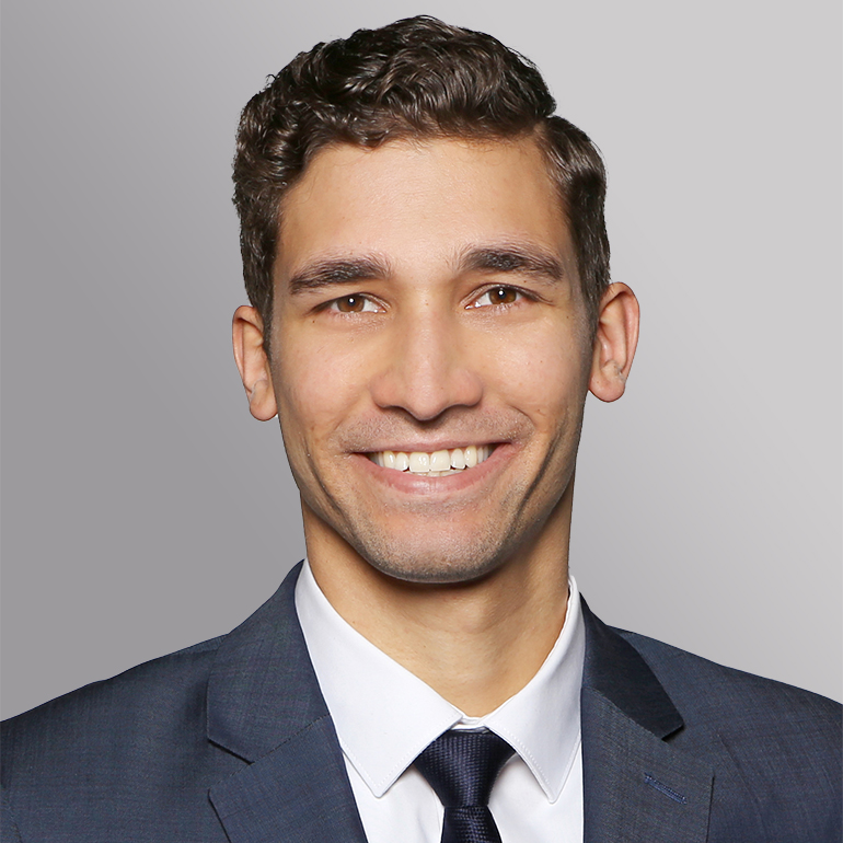 Ein junger Mann mit braunen, lockigen Haaren lächelt freundlich in die Kamera. Er trägt einen dunkelblauen Anzug mit einer weißen Hemdbluse und einer dunkelblauen Krawatte. Der Hintergrund ist grau und neutral, wodurch sein Gesicht hervortritt.