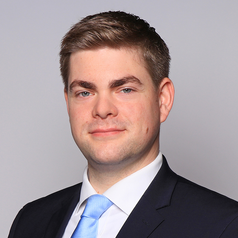 Ein lächelnder Mann mit kurzen, blonden Haaren trägt einen Anzug und eine hellblaue Krawatte. Der Hintergrund ist grau. Die Aufnahme zeigt seinen Kopf und Oberkörper.