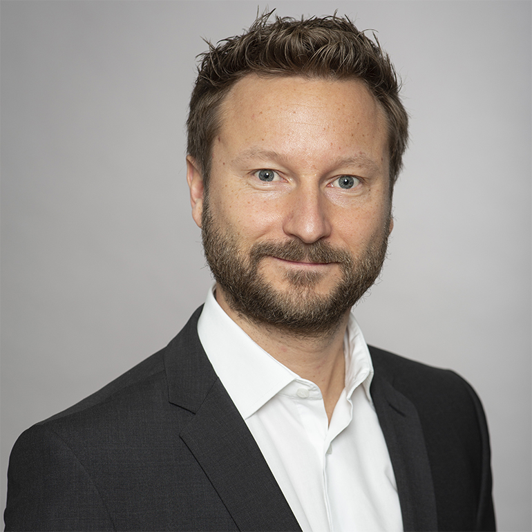 Ein Mann mit einem leichten Bart trägt einen Anzug und ein weißes Hemd. Er schaut freundlich in die Kamera, mit einem neutralen Hintergrund. Seine Haare sind kurz und leicht gewellt.