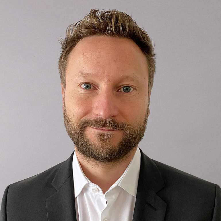 Ein Mann mit einem kurzen, welligen Bart und braunen Augen lächelt freundlich in die Kamera. Er trägt einen schwarzen Anzug und ein weißes Hemd. Der Hintergrund ist grau.