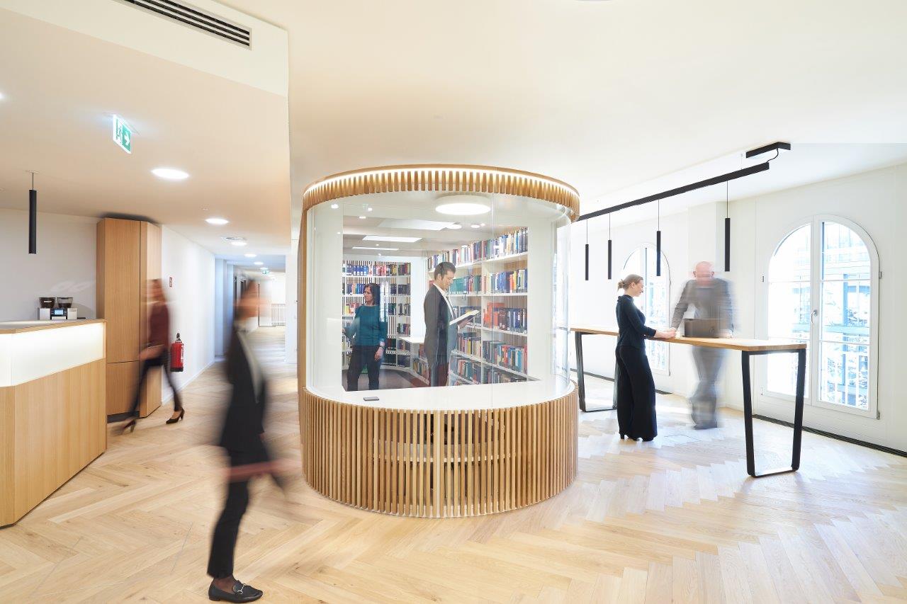 Ein moderner Raum mit einem runden, gläsernen Bücherregal in der Mitte. Mehrere Personen stehen oder bewegen sich darin. An einer langen Holztheke interagiert eine Frau mit einem Mann, der vorbeikommt. Der Boden ist aus hellem Holz, und große Fenster lassen viel Licht herein.