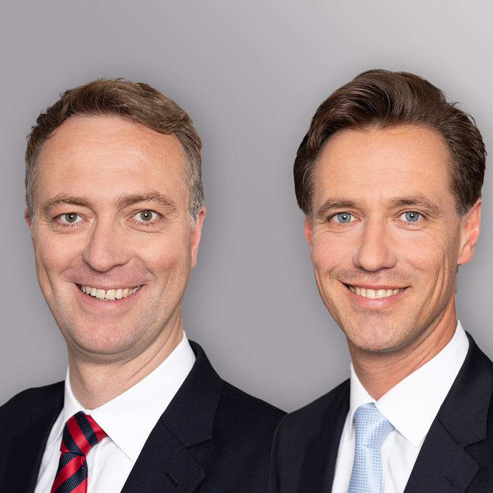 Zwei Männer in Anzügen stehen nebeneinander und lächeln freundlich in die Kamera. Der linke Mann trägt eine Krawatte mit rotem und blauem Muster, der rechte eine hellblaue Krawatte. Der Hintergrund ist neutral grau.