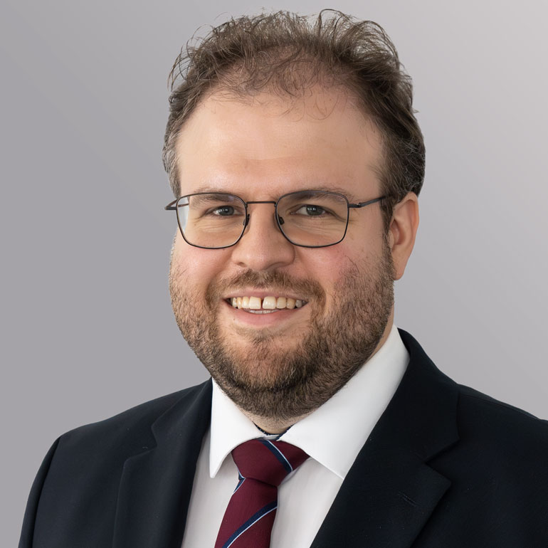 Ein lächelnder Mann mit Brille und einem kurzen Bart trägt einen Anzug mit einem roten und blauen gestreiften Krawatte. Der Hintergrund ist grau.