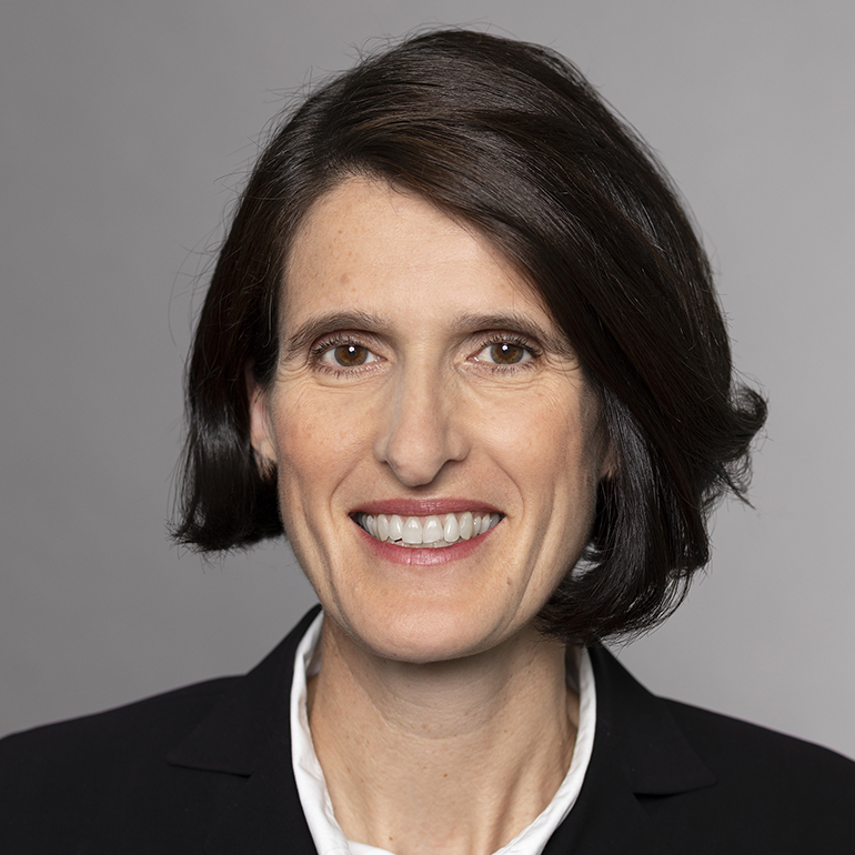 Porträt einer lächelnden Frau mit schulterlangen, dunklen Haaren, die einen schwarzen Blazer und ein weißes Hemd trägt. Der Hintergrund ist grau, was die Gesichtszüge der Frau betont.