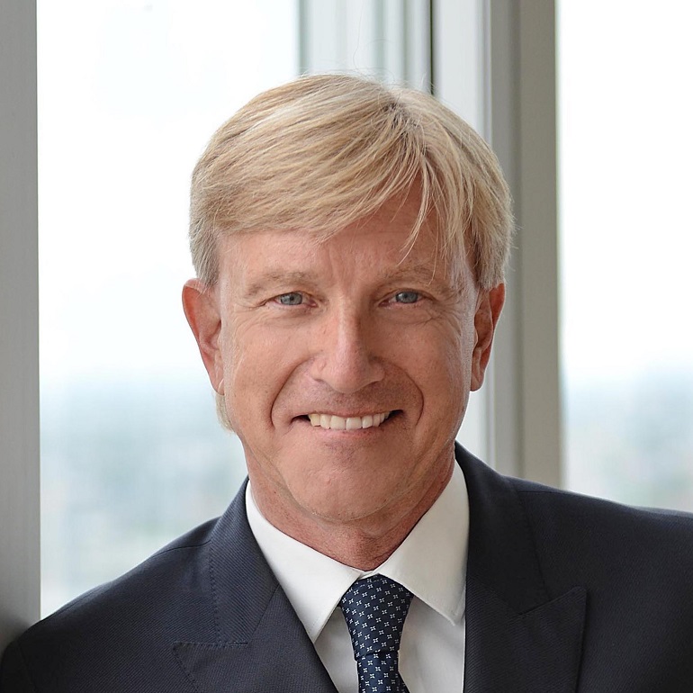 Ein lächelnder Mann mit blondem Haar und einem Anzug steht vor einem Fenster. Er hat eine freundliche Ausstrahlung und trägt eine Krawatte mit einem Muster. Der Hintergrund ist hell und unscharf.