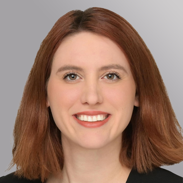 Porträt einer Frau mit schulterlangen, rotbraunen Haaren und einem freundlichen Lächeln. Sie hat eine helle Hautfarbe und trägt ein schwarzes Oberteil. Der Hintergrund ist graufarben.