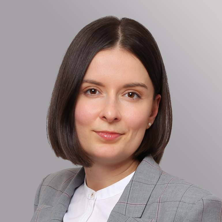 Portrait einer jungen Frau mit schulterlangen, glatten Haaren und einem neutralen Ausdruck. Sie trägt ein graues Jackett mit karierten Mustern und ein weißes Hemd. Der Hintergrund ist einfarbig grau.