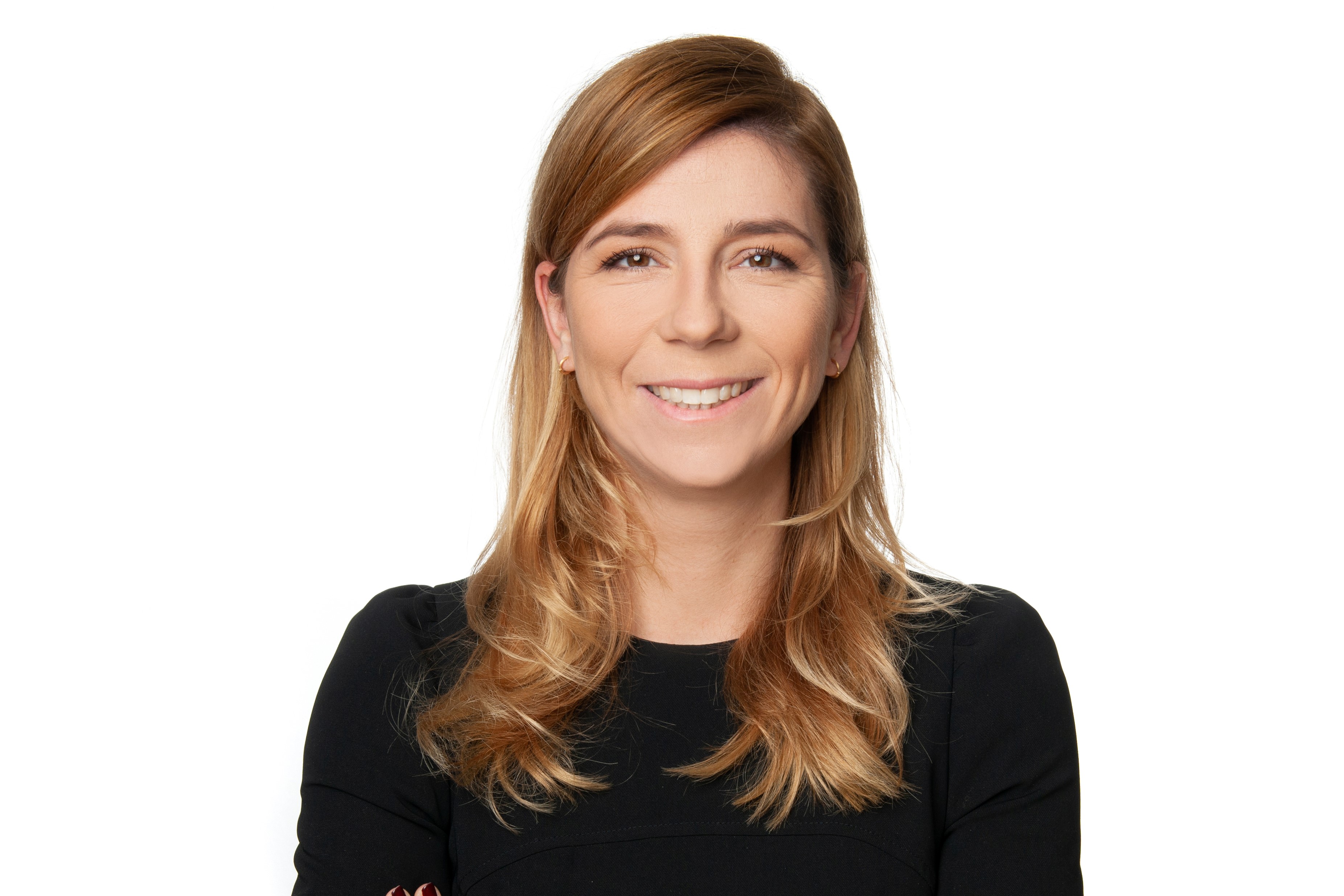 Porträt einer lächelnden Frau mit langen, blonden Haaren, die in einem schwarzen Oberteil gekleidet ist. Sie steht mit verschränkten Armen vor einem neutralen, weißen Hintergrund. Ihr Gesichtsausdruck strahlt Freundlichkeit und Selbstbewusstsein aus.