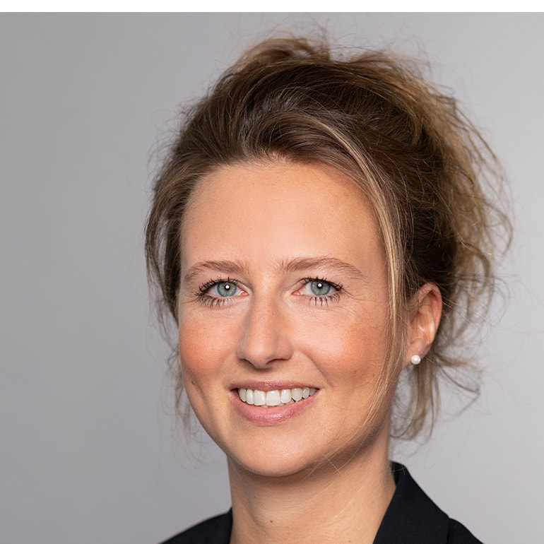 Porträt einer lächelnden Frau mit blonden, lockigen Haaren. Sie trägt einen schwarzen Blazer und hat Ohrstecker. Der Hintergrund ist grau und neutral gehalten. Die Frau hat einen freundlichen und selbstbewussten Ausdruck.