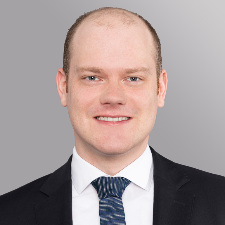 Ein junger Mann mit kurzen, hellen Haaren lächelt in die Kamera. Er trägt einen dunklen Anzug mit einem weißen Hemd und einer gemusterten Krawatte. Der Hintergrund ist einfarbig grau.