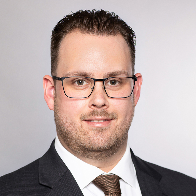 Ein Mann mit Brille trägt einen dunklen Anzug und eine helle Krawatte. Er hat kurzes, dunkles Haar und lächelt in die Kamera. Der Hintergrund ist hell und einfarbig.