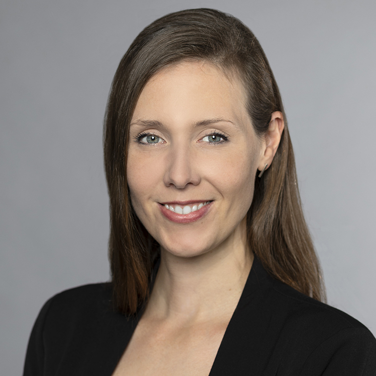 Porträt einer lächelnden Frau mit langen, braunen Haaren, die einen schwarzen Blazer trägt. Der Hintergrund ist neutral und grau, was den Fokus auf ihr freundliches Gesicht und ihre Ausstrahlung lenkt. Sie sieht freundlich und professionell aus.