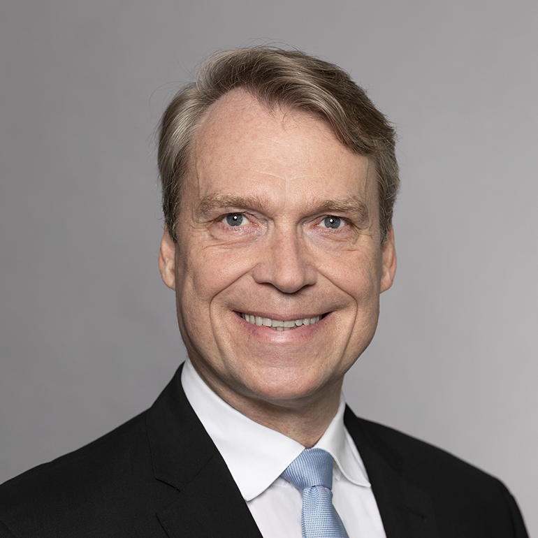 Ein Mittelaltermann mit kurzen, blonden Haaren und einem freundlichen Lächeln trägt einen schwarzen Anzug und eine hellblaue Krawatte. Der Hintergrund ist grau.