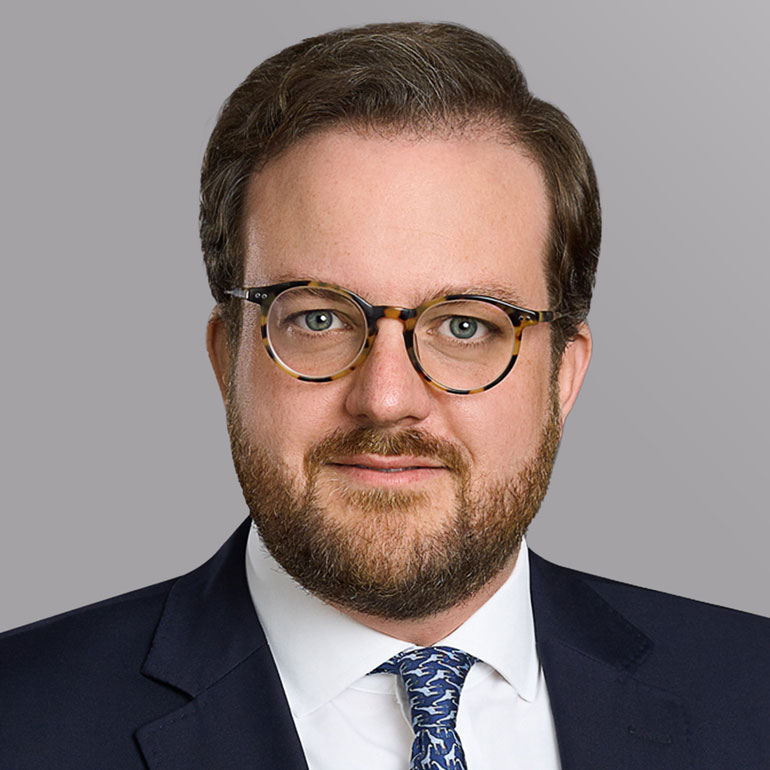 Ein Mann mit Brille und Bart trägt einen dunklen Anzug und ein helles Hemd mit einer gemusterten Krawatte. Er blickt direkt in die Kamera vor einem grauen Hintergrund.