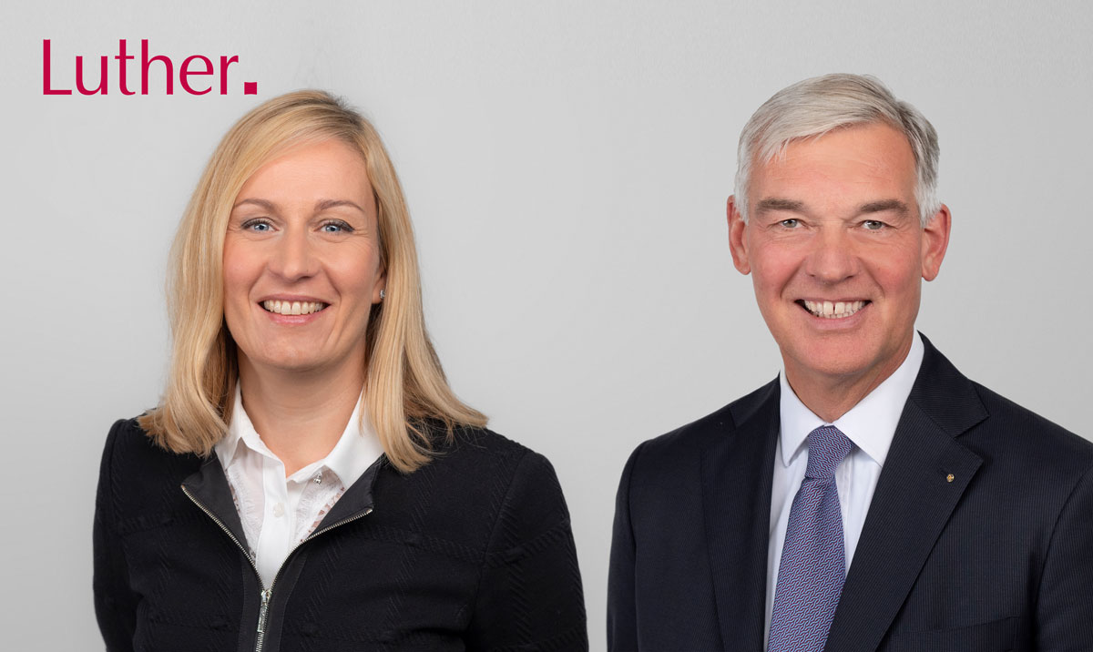A smiling man in a suit and a woman in a black jacket and white shirt pose together against a light gray background. The text “Luther.” appears in the top left corner.