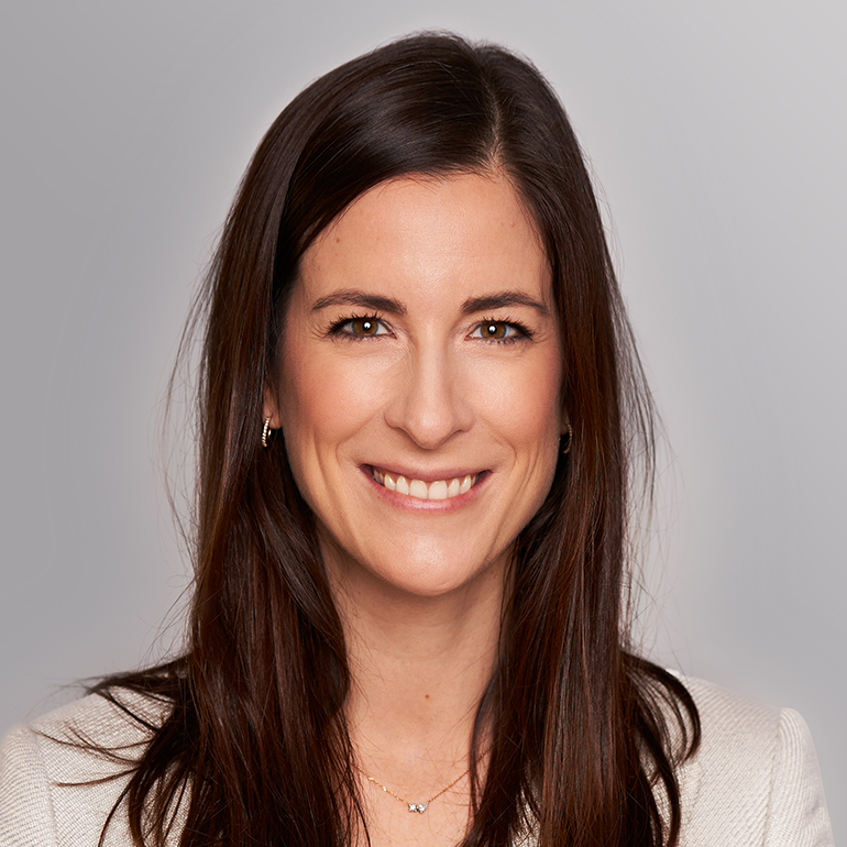 Porträt einer lächelnden Frau mit langen, dunklen Haaren. Sie trägt eine helle Jacke und eine zarte Halskette. Der Hintergrund ist in neutralen Grautönen gehalten.