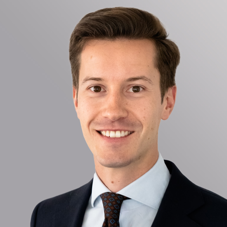 Ein junger Mann mit kurzer, brauner Haarfrisur lächelt in die Kamera. Er trägt ein dunkles Sakko und ein hellblaues Hemd mit einer Krawatte in Braun mit Muster. Der Hintergrund ist einfarbig grau.